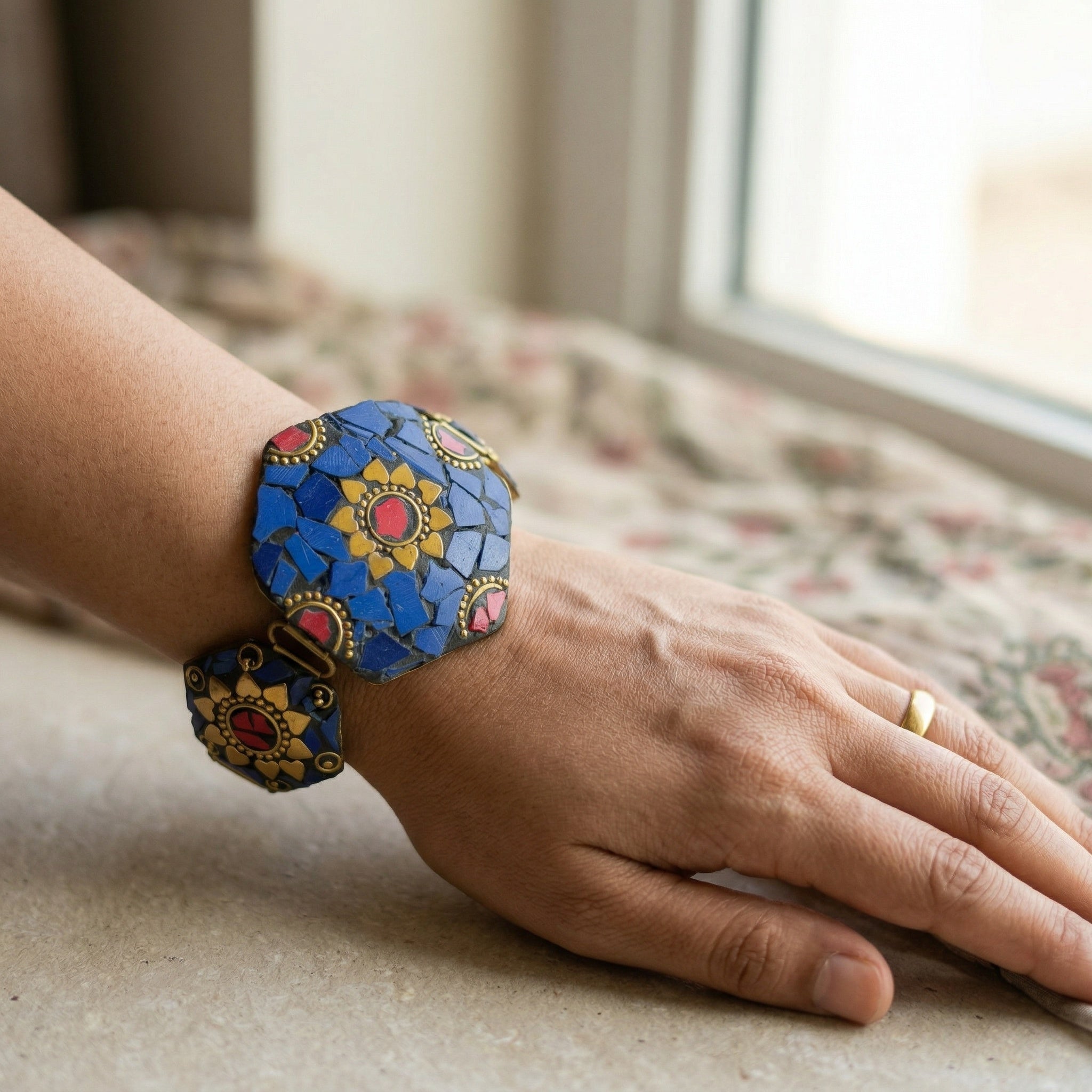 Hand wearing a colorful bracelet with a floral design, sitting on a surface with a window in the background.