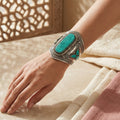 Hand wearing a silver cuff with turquoise stones on a textured surface.