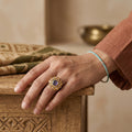 Hand wearing a gold ring with a purple gemstone on a wooden surface.