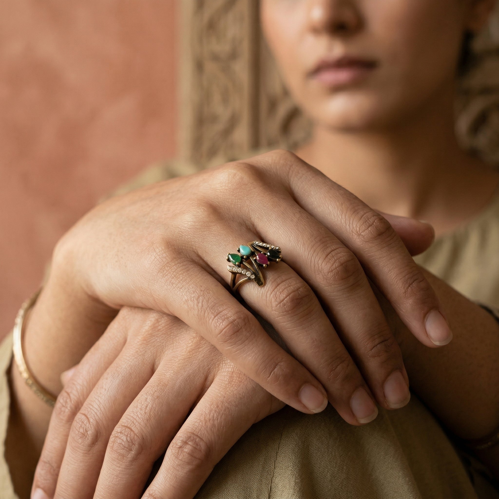 Close-up of a person's hands with a ring featuring green and red gemstones, wearing a beige outfit.