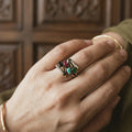 Hand wearing a ring with colorful gemstones against a wooden background