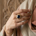Hand wearing a gold ring with a blue gemstone against a textured beige background