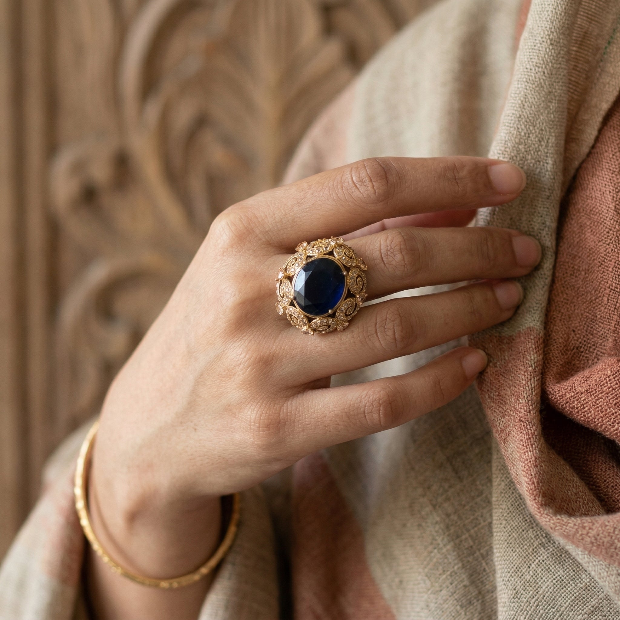 Hand wearing a gold ring with a blue gemstone against a textured beige background
