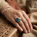 Hand wearing a ring with blue, green, and beige stones on a wooden surface.