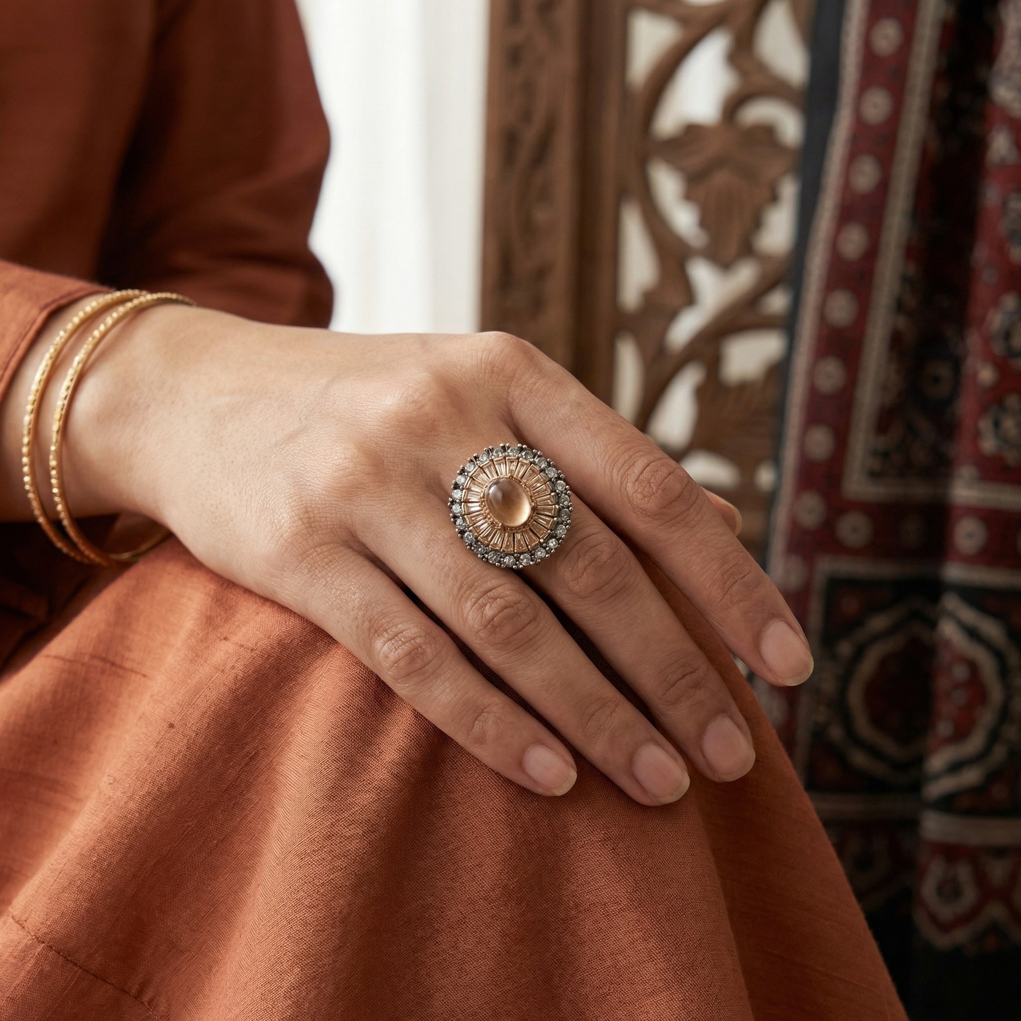 Hand wearing a decorative ring with intricate design, set against a patterned fabric background.