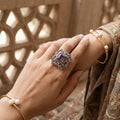 Hand wearing a decorative ring with a purple gemstone and gold bracelet against a blurred background