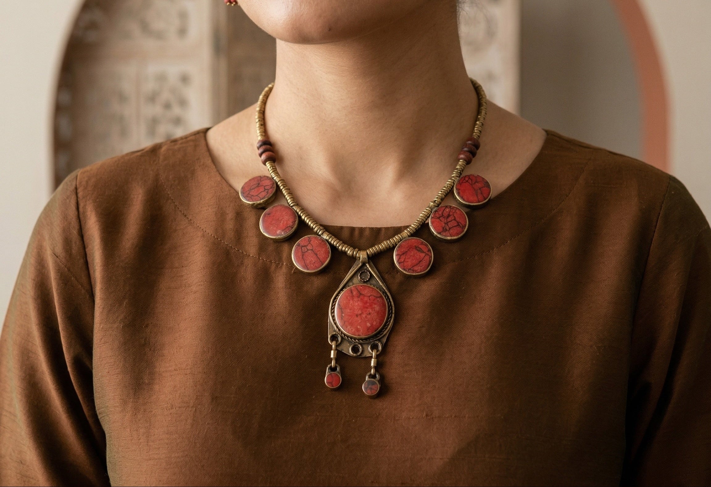 Person wearing a detailed necklace with red stones and brown top.